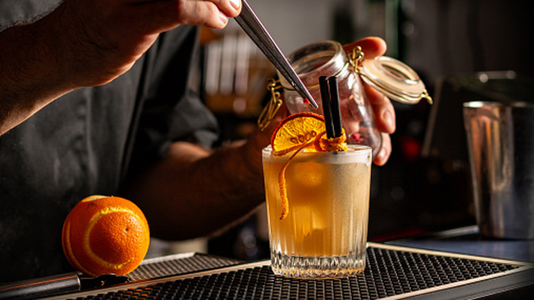 Bartender preparing smoky spiced orange cocktail decorated with dehydrated orange slices and fresh orange peel.
