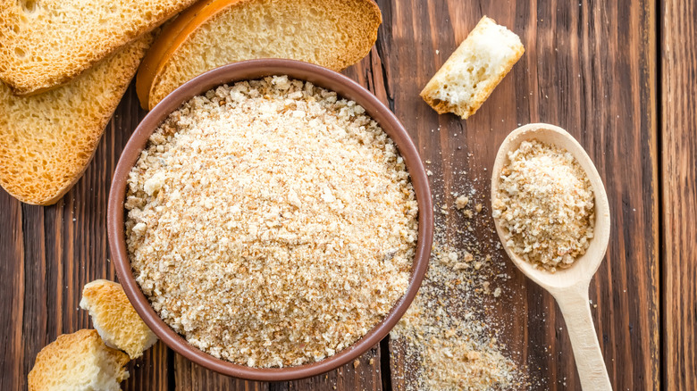 A bowl and spoon filled with breadcrumbs and surrounded by bread pieces