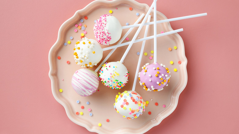 Various white and pink frosted and sprinkled cake pops on a plate and pink background