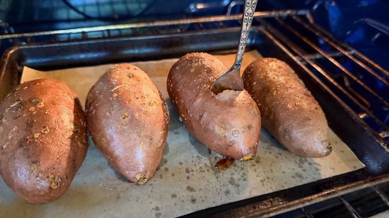 Testing baked sweet potatoes on parchment-lined baking sheet with fork in oven