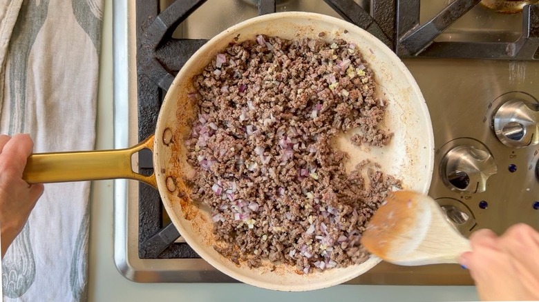 Stirring ground beef with red onion and garlic using wooden spoon in white skillet on stovetop