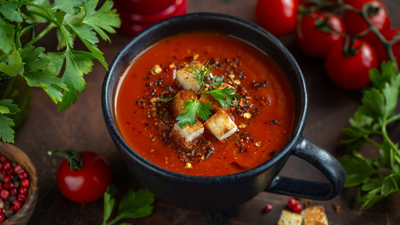 Tomato soup in a black mug, surrounded by whole tomatoes and herbs
