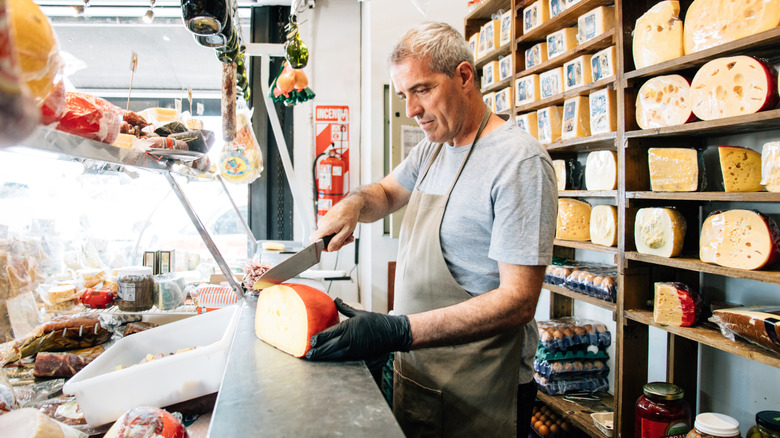 A man cuts into a wheel of cheese behind a deli counter