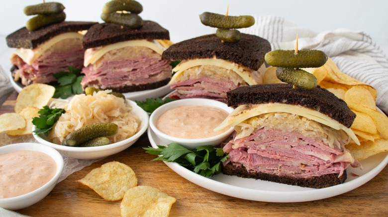 sliced reuben sandwiches with pickles on plates surrounded by bowls of dressing