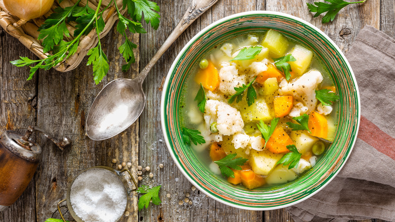 Bowl of vegetable soup on a wooden board, fresh herbs and a spoon on the side