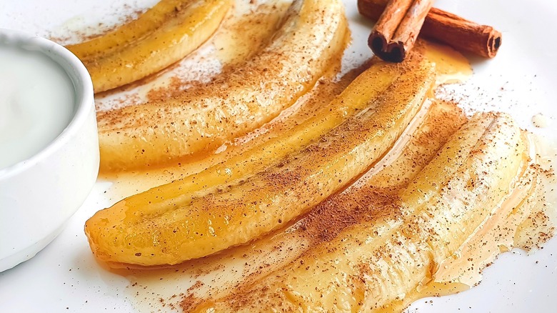 Halved caramelized bananas topped with cinnamon next to a small bowl of yogurt and two whole cinnamon sticks.