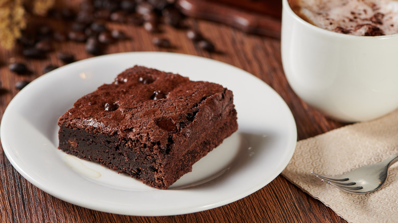 Chocolate brownie on a white plate next to a cappuccino