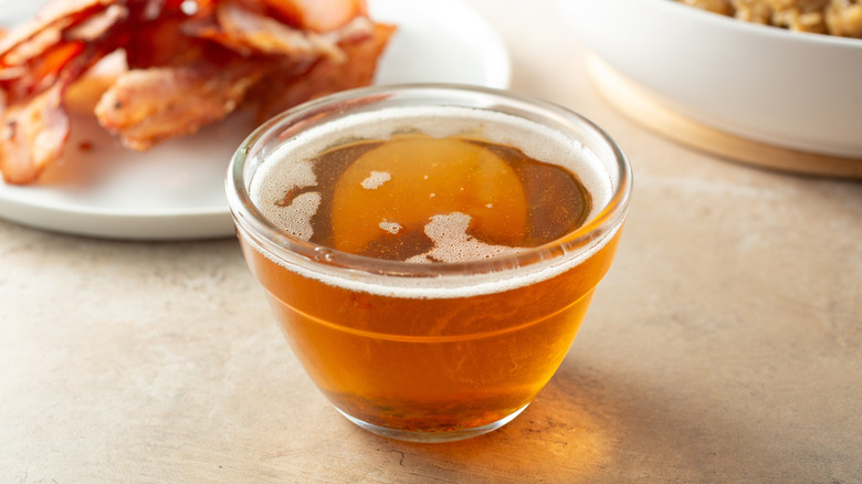 A small bowl of bacon grease in front of a plate of bacon.