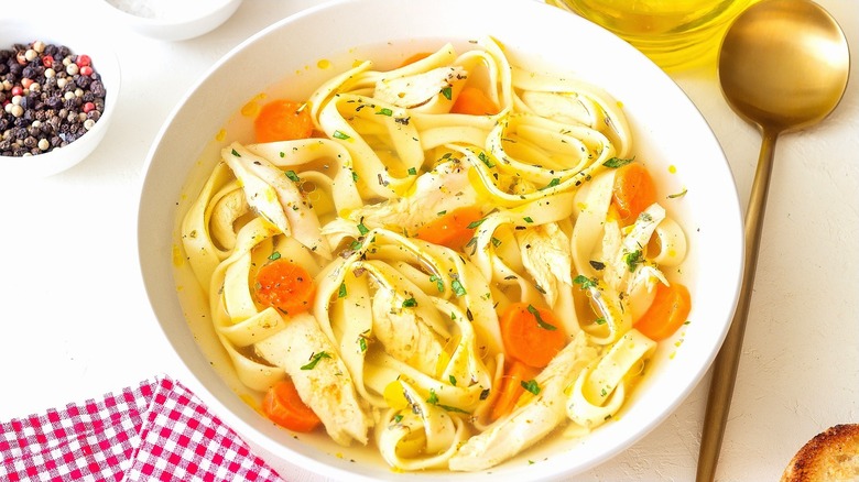 Chicken noodle soup in a white bowl next to a gold spoon and small bowl of whole peppercorns