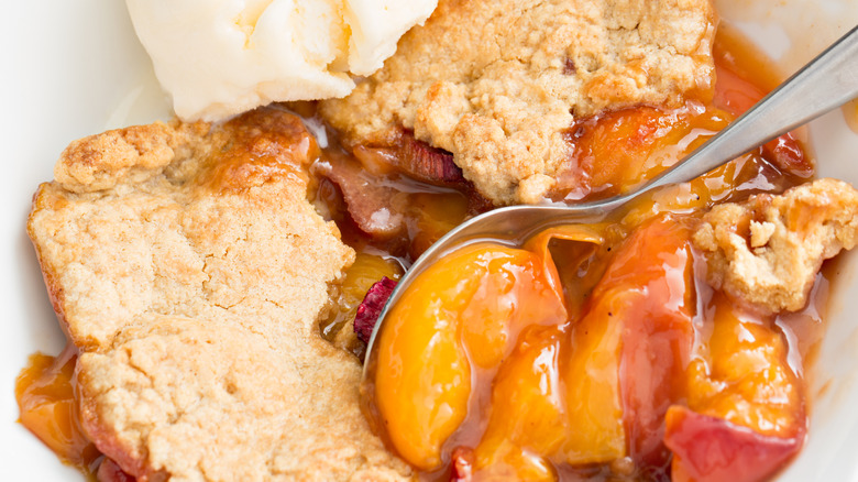 An overhead close-up of a peach cobbler with a scoop of vanilla ice cream in a white bowl.
