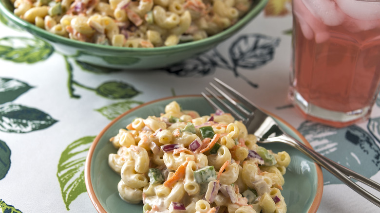 Green plate of homemade macaroni salad with a fork next to a drink and serving bowl of it, all on a leafy tablecloth