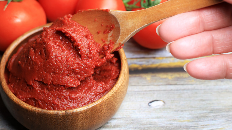A hand scooping out tomato paste with a wooden spoon