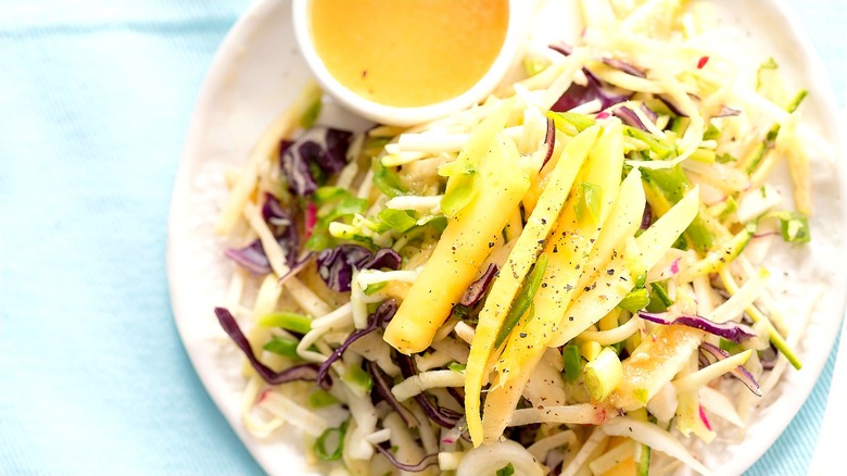 Mango-infused coleslaw next to a small cup of salad dressing on a cream-colored plate