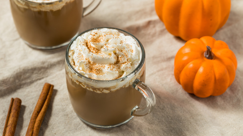 Pumpkin spice latte in a glass mug with whipped cream with cinnamon sticks and pumpkins in the background