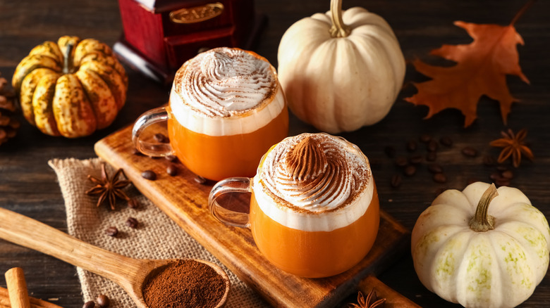 Two pumpkin spice lattes with whipped cream on a wooden board with fall decor in the background