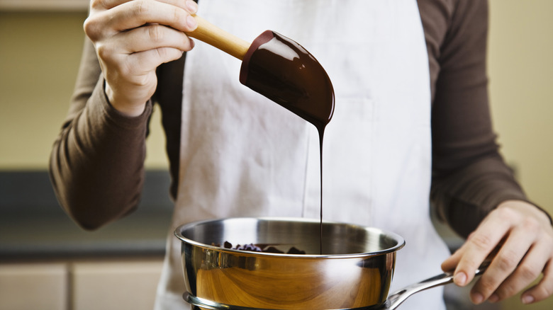 A person holds a spatula dripping with melted chocolate over a metal bowl