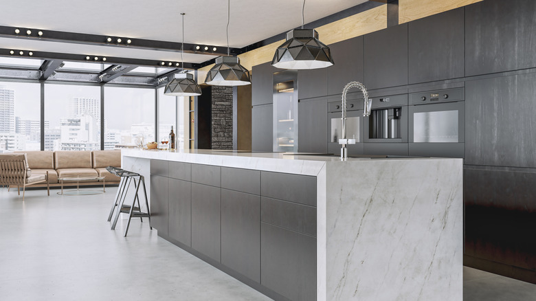 A marble island in a modern kitchen with floor-to-ceiling windows, hidden cabinets, and low-hanging light fittings
