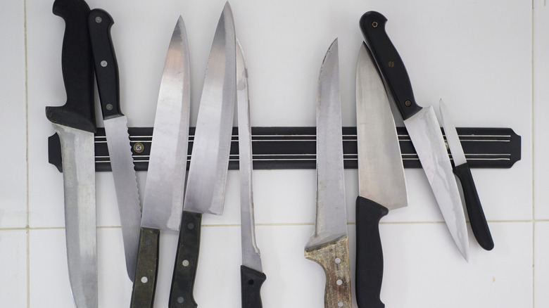 A collection of kitchen knives on a magnetic strip attached to a white tiled wall