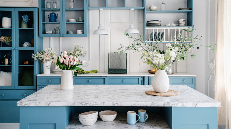 A marble countertop with blue painted shelves and cabinets in the background with two vases of white flowers on top