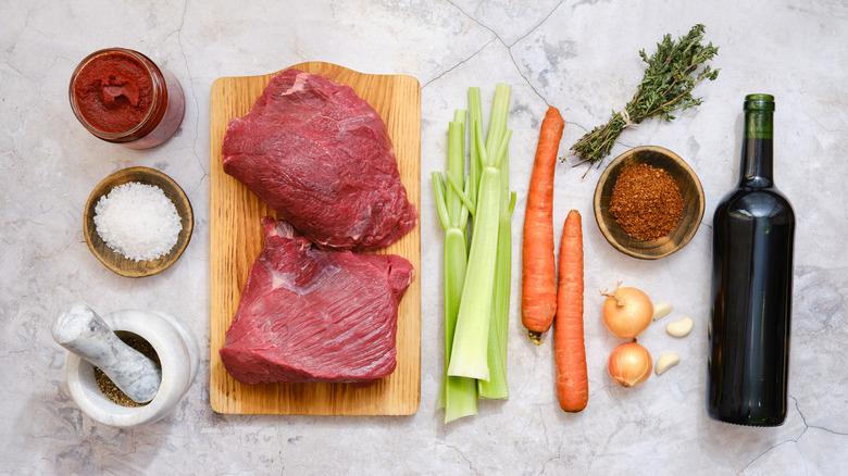 Slabs of red meat on a wooden cutting board next to spices, a jar of tomato paste, fresh vegetables, and a bottle of red wine, all on a white marble countertop