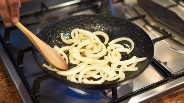 Hand holding wooden spoon stirring onions sautéing in a frying pan.
