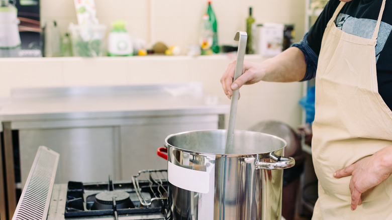 A person is seen stirring the contents of a stockpot on a stove with a long utensil