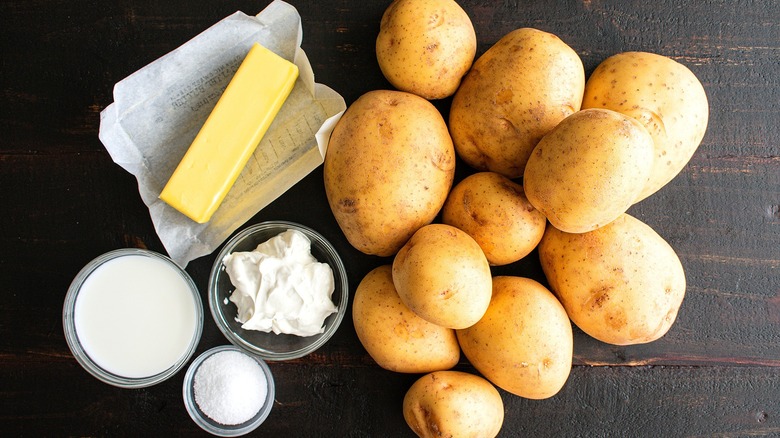 Unpeeled yellow potatoes next to an unwrapped stick of butter and small bowls of milk, sour cream, and salt