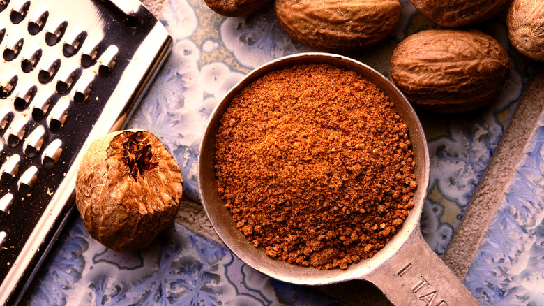 Ground nutmeg in a metal tablespoon with whole nutmeg and a grater nearby on a tile countertop.