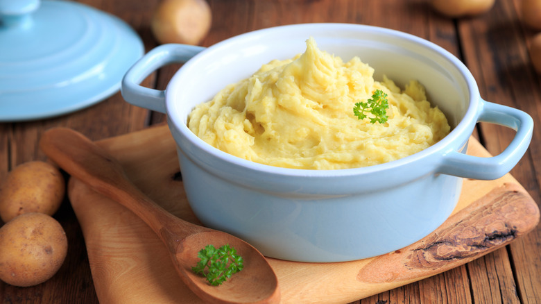 Homemade mashed potatoes in a blue pot on a wooden serving platter with a wooden spoon with herbs, potatoes, and a matching blue lid on a wooden table.