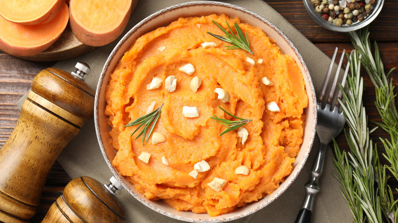 Bowl of mashed sweet potatoes topped with herbs and nuts on a table with salt and pepper shakes, a fork, and herbs