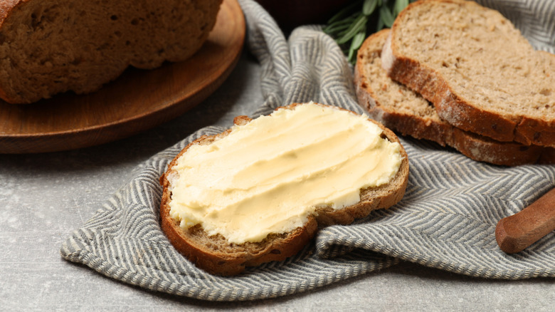 A slice of fresh, whole wheat bread covered in fresh, soft butter, other slices of bread and the remaining loaf behind.