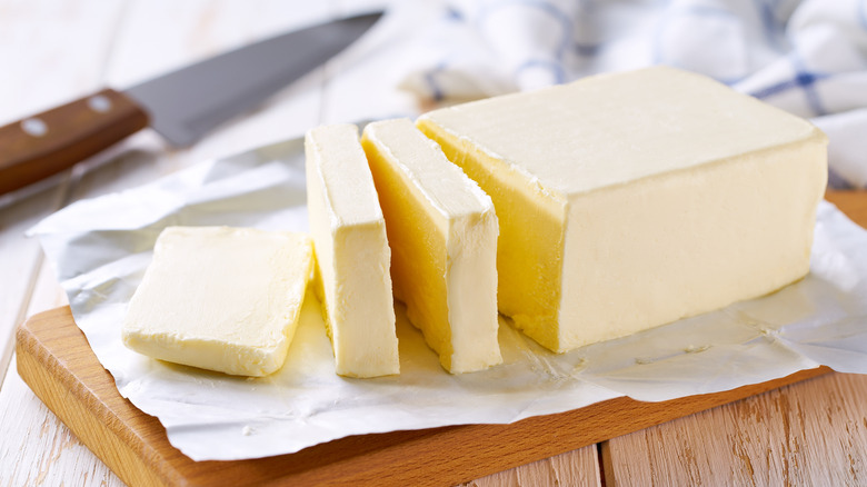 White, slightly yellow, creamy butter sliced into pats on top of a wooden cutting board over a wooden table, a cheese/butter knife in the backround.