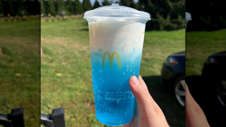 Person's hand holding a blue, white foam-topped McDonald's Sprite Berry Blast out against a background of trees and grass