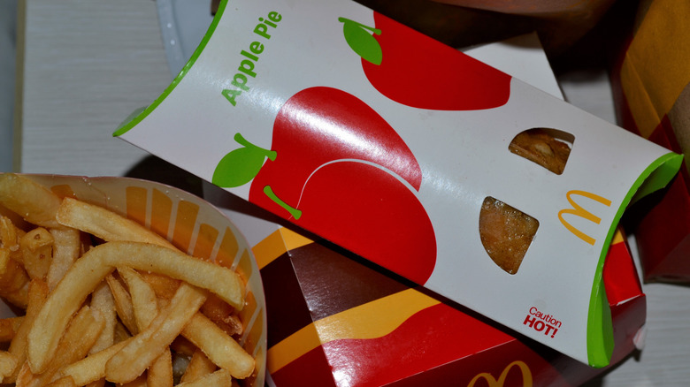 A McDonald's apple pie in its white, green, and red cardboard sleeve sits on top of a closed box of food and beside an open container of French fries on a tabletop.