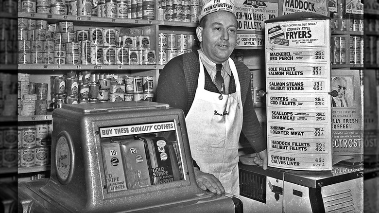 Black and white photo of a man standing at a grocery store counter in front of a coffee bean machine and a pricing list for seafood.
