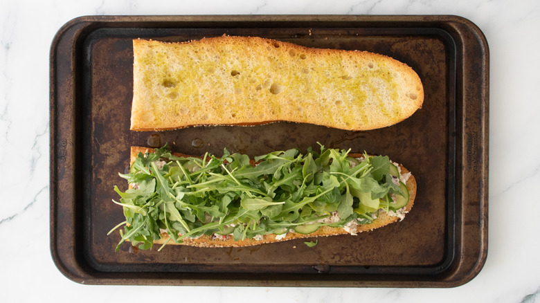 A layered tuna salad on a large toasted baguette topped with baby arugula