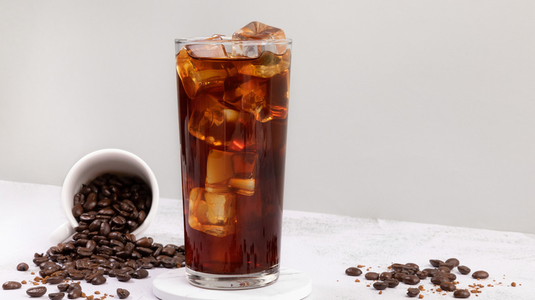 A glass of iced coffee on a table with coffee beans pouring out from a white mug.