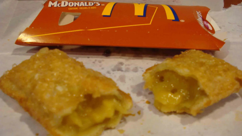 A McDonald's fried apple pie, split open to reveal the filling, sits on a paper covering on a table beside its red cardboard packing sleeve.