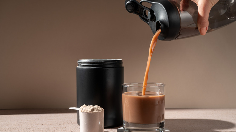 Hand holding tumbler with chocolate drink pouring it into a short glass with scoop of powder beside it.