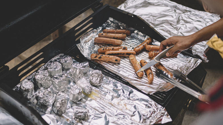 Person tending aluminum foil lined grill grates with various foods wrapped in foil over hot coals.