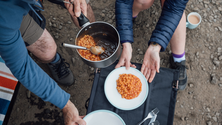 Two people serving a bot of baked beans on a campsite into white bowl.