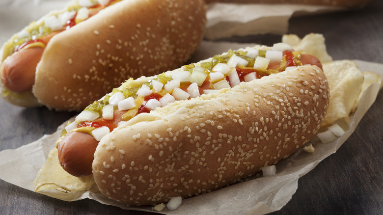 Two hot dogs in sesame buns on baking paper on table.