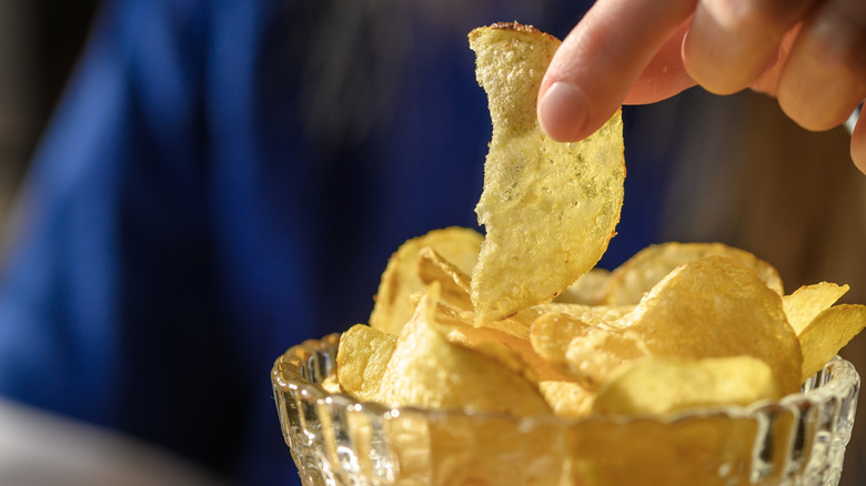 Hand reaching into glass bowl of potato chips.