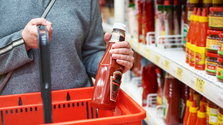Shopper holding bottle of ketchup as they put it into shopping basket in store.