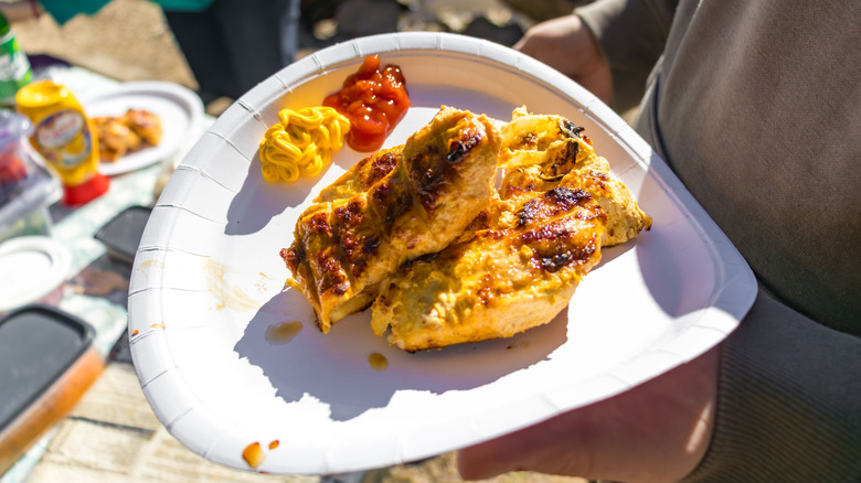 A couple of pieces of grilled chicken on white paper plate with condiments on side.