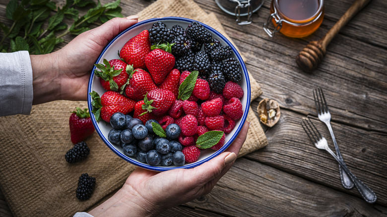 Woman holding bowl of fresh blueberries, strawberries, and blackberries over wood table.