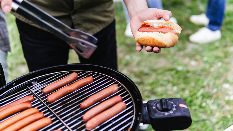 Person grilling hot dogs with tongs in hand in grassy area.