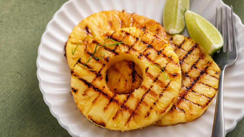 Grilled pineapple rings on white plate next to fresh limes with fork on green counter.
