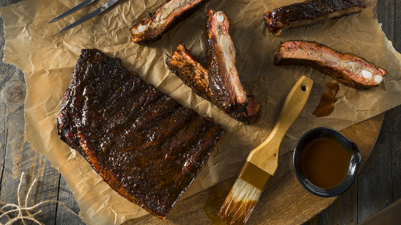 Glaze-covered rack of ribs on baking paper resting over wooden cutting board on countertop.