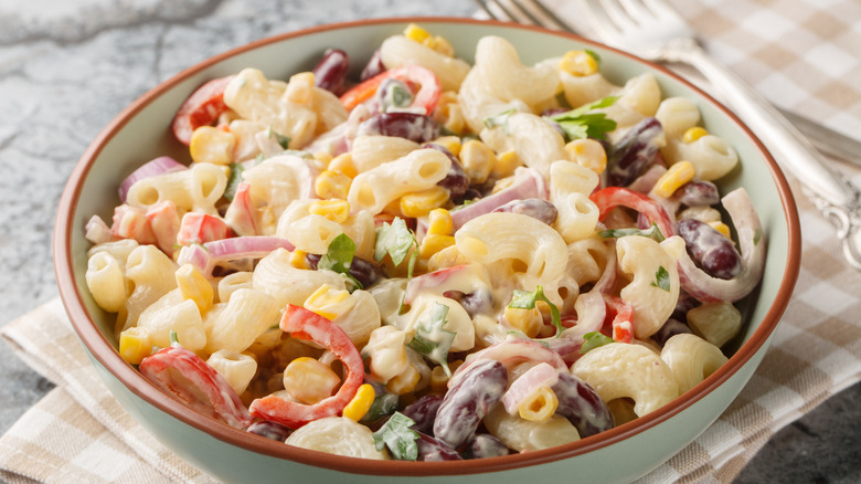 Macaroni salad with cabbage, onions, peppers, and corn in ceramic bowl on tea towel next to fork.